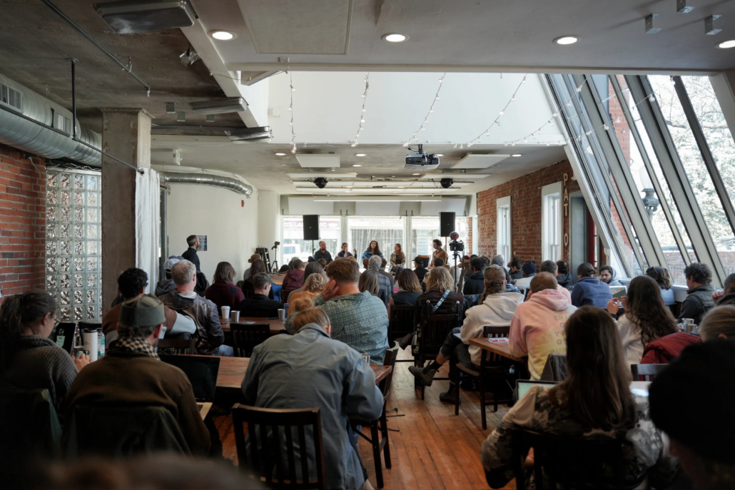 Full event setup in the Agora Room at Boulder Riverside with audience seated at long farm tables facing a panel