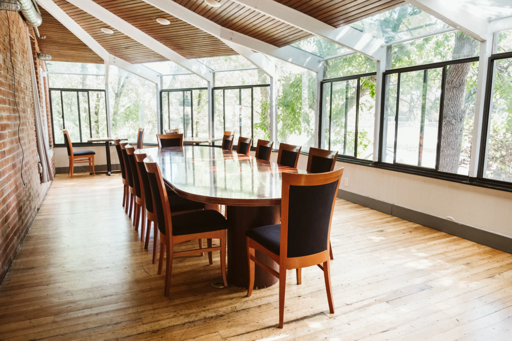 Creek Room private meeting and event space at Boulder Riverside with floor-to-ceiling windows, wood-slat ceiling, oval conference table, and tree-lined views overlooking Boulder Creek, seats up to 20 guests